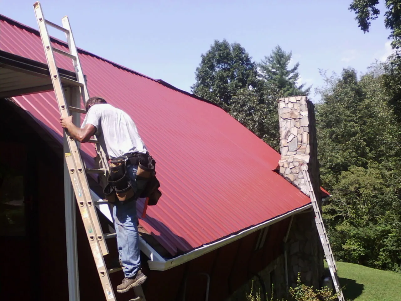 Roofer on a ladder beside a red metal roof and stone chimney — Soffit & Fascia Repair by Roofing Brothers in Alburtis